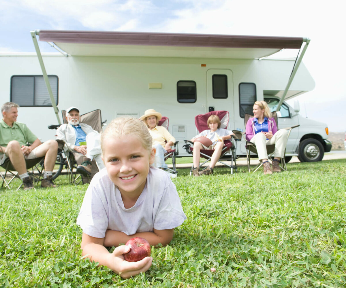 Girl in the grass holding an apple while family talks near RV.