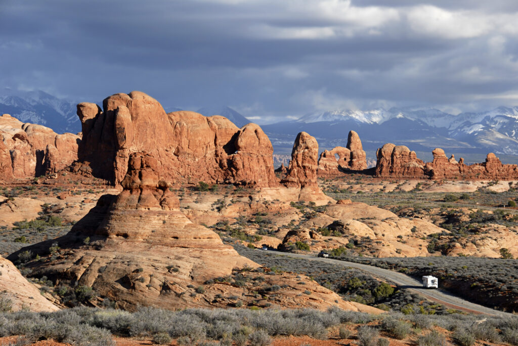 A distant RV drives on a road with large rock formations and mountains in the background