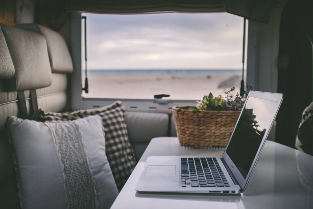 A laptop is open on a table inside of an RV with decorative pillows, a basket, and a window.