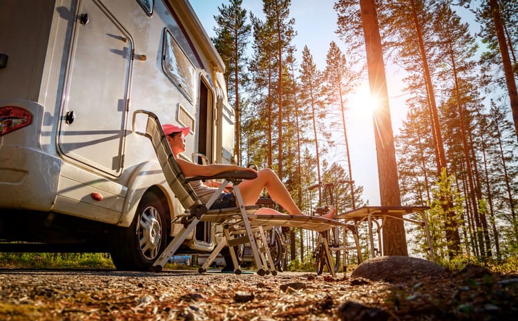 Woman reclining in a chair, in a forest, next to her RV camper.