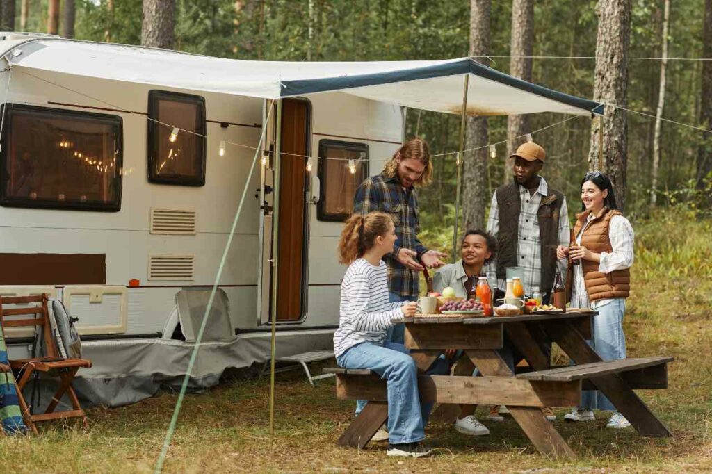 A group of friends sit on a picnic bench outside of an RV with a tent and lights