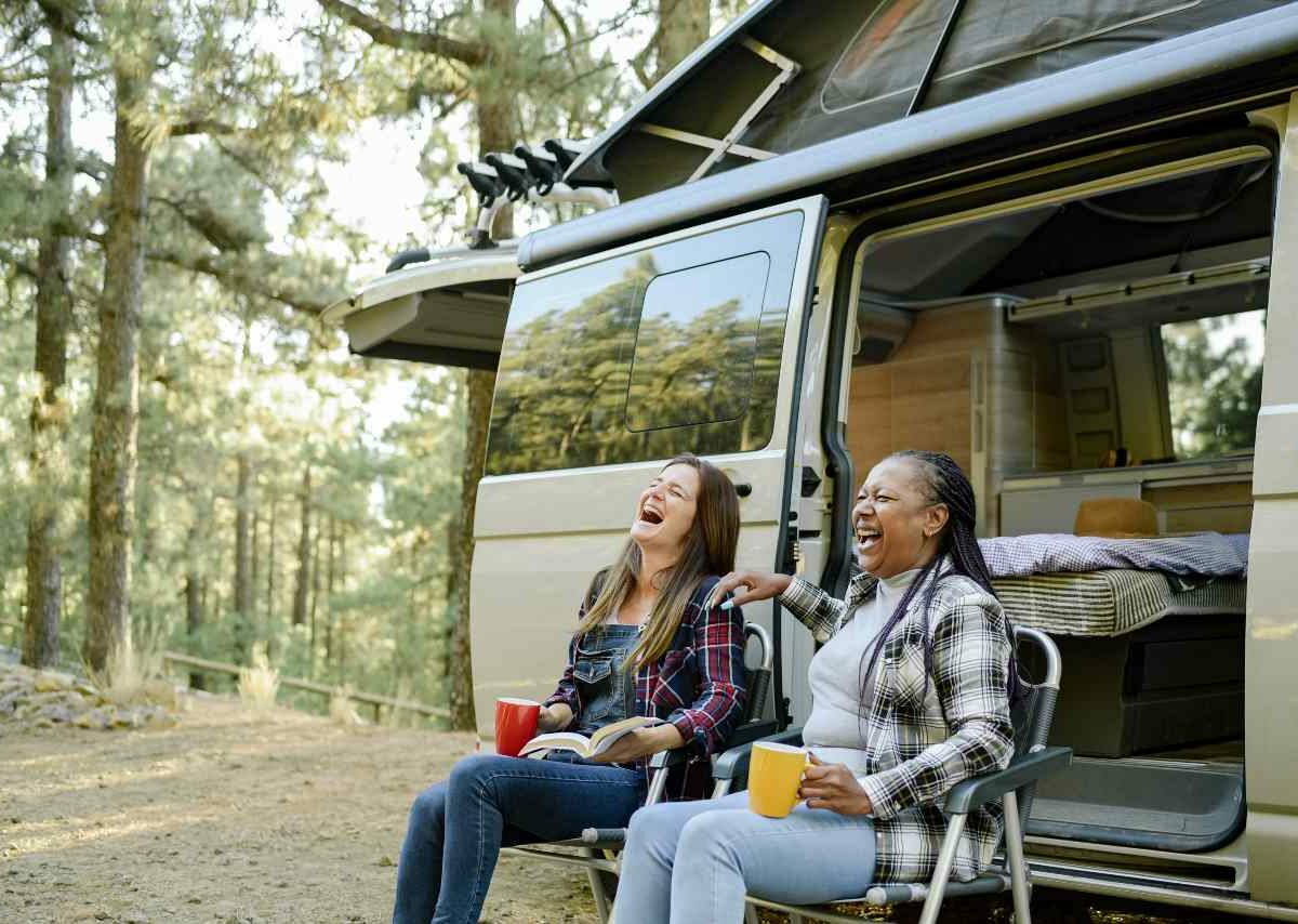 Two friends sit in chairs and chat outside of a small RV