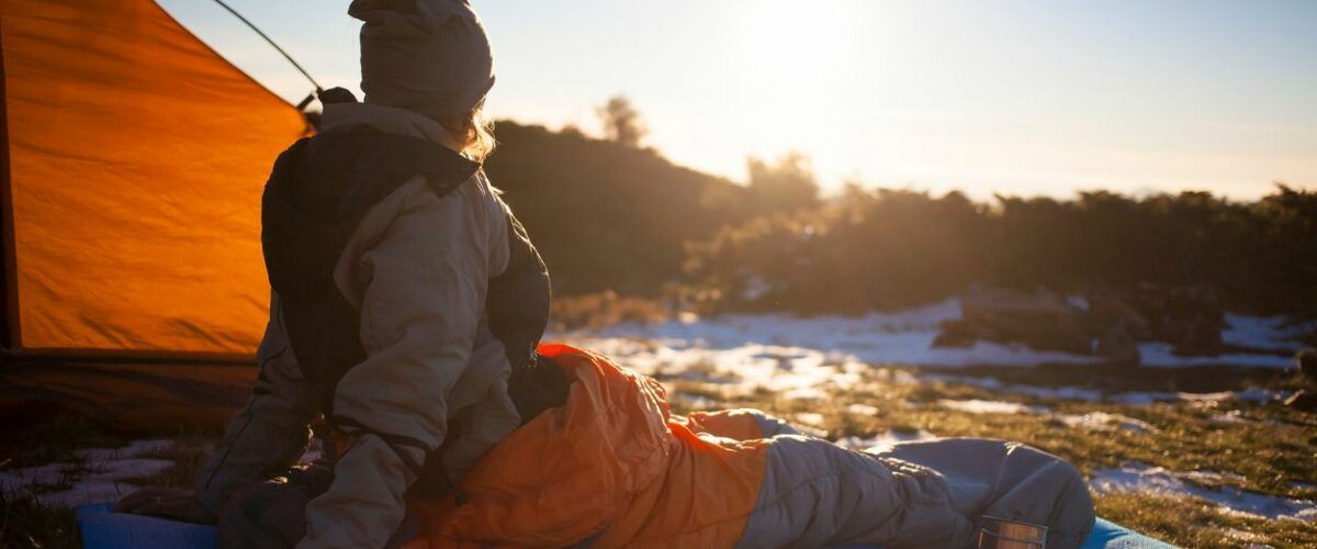 A person sits inside a camping bag on a mat and looks out into a snowy nature landscape as the sun rises.
