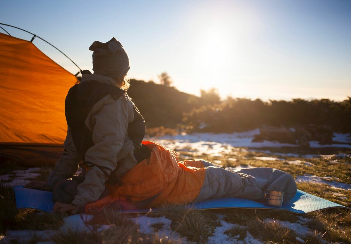 A person sits inside a camping bag on a mat and looks out into a snowy nature landscape as the sun rises.