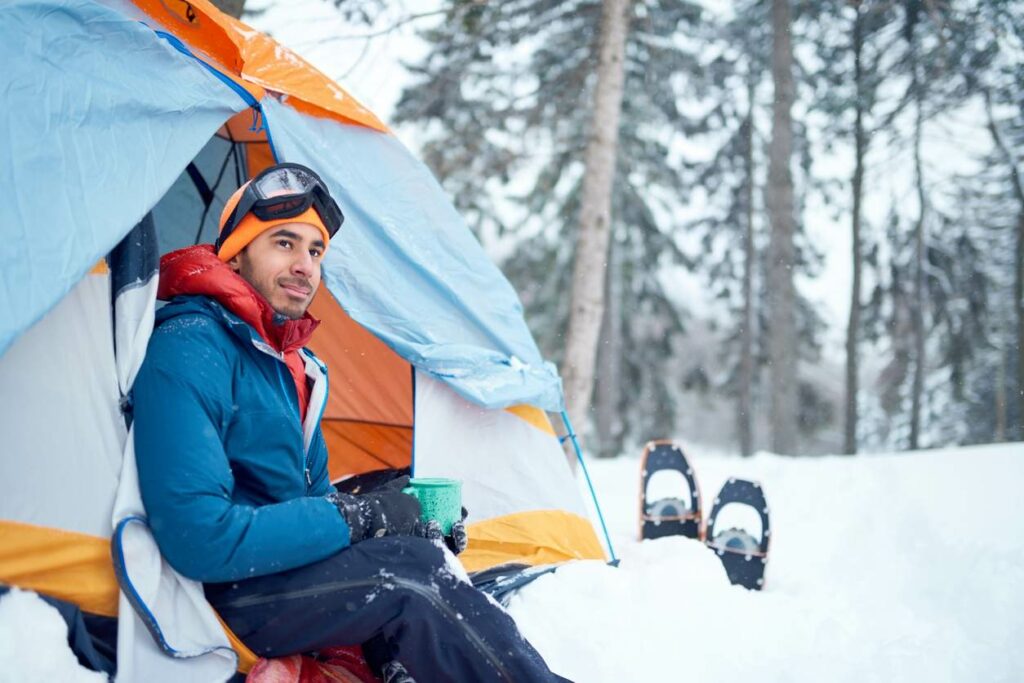A man sits in the opening of a tent surrounded by snow while drinking out of a coffee cup