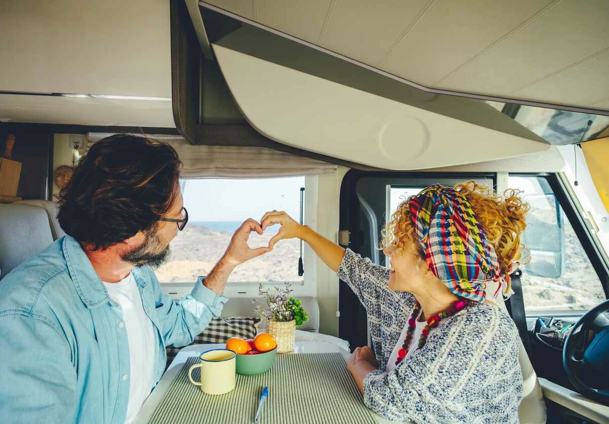 A man and woman sit in an RV and use their hands to form a heart shape in front of the window.