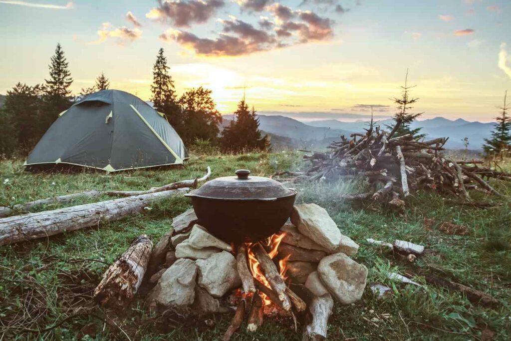 A large pot is set on a campfire supported by stones with a nearby tent and pile of branches