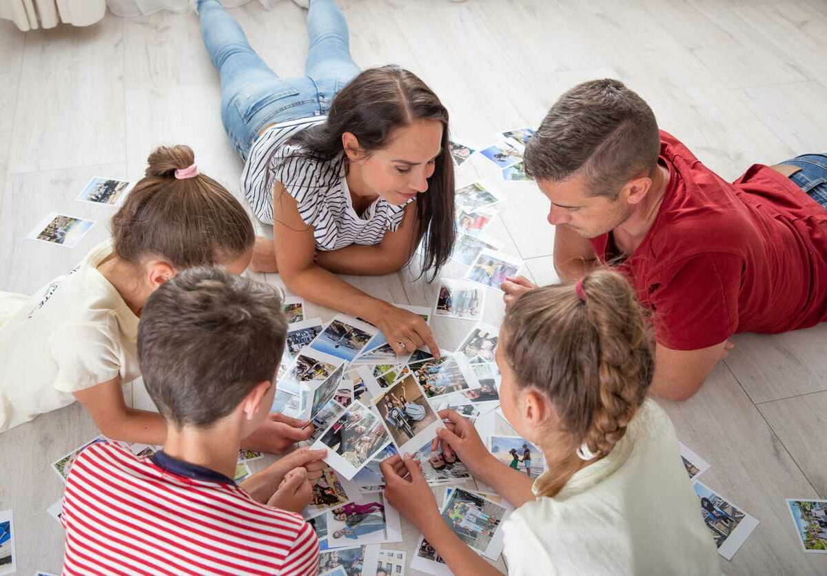 A family of parents and three children lay on a floor around a spread of photos looking at and pointing to them