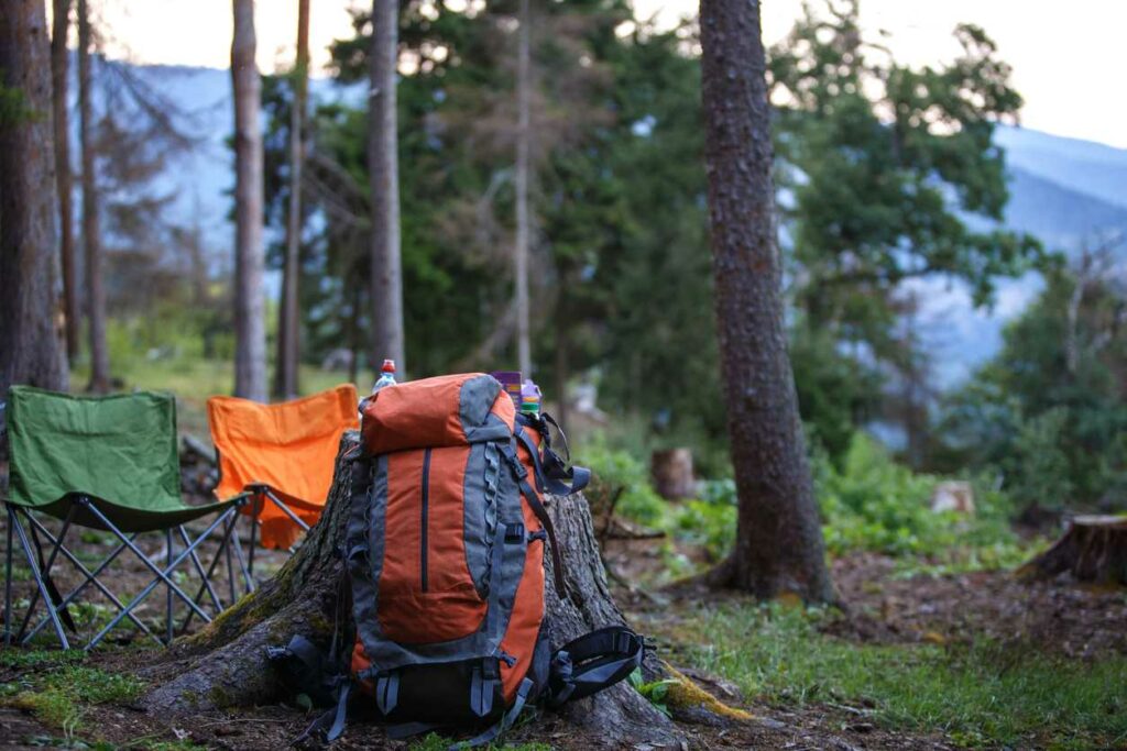 A large hiking backpack rests against a tree trump alongside folding chairs in the forest