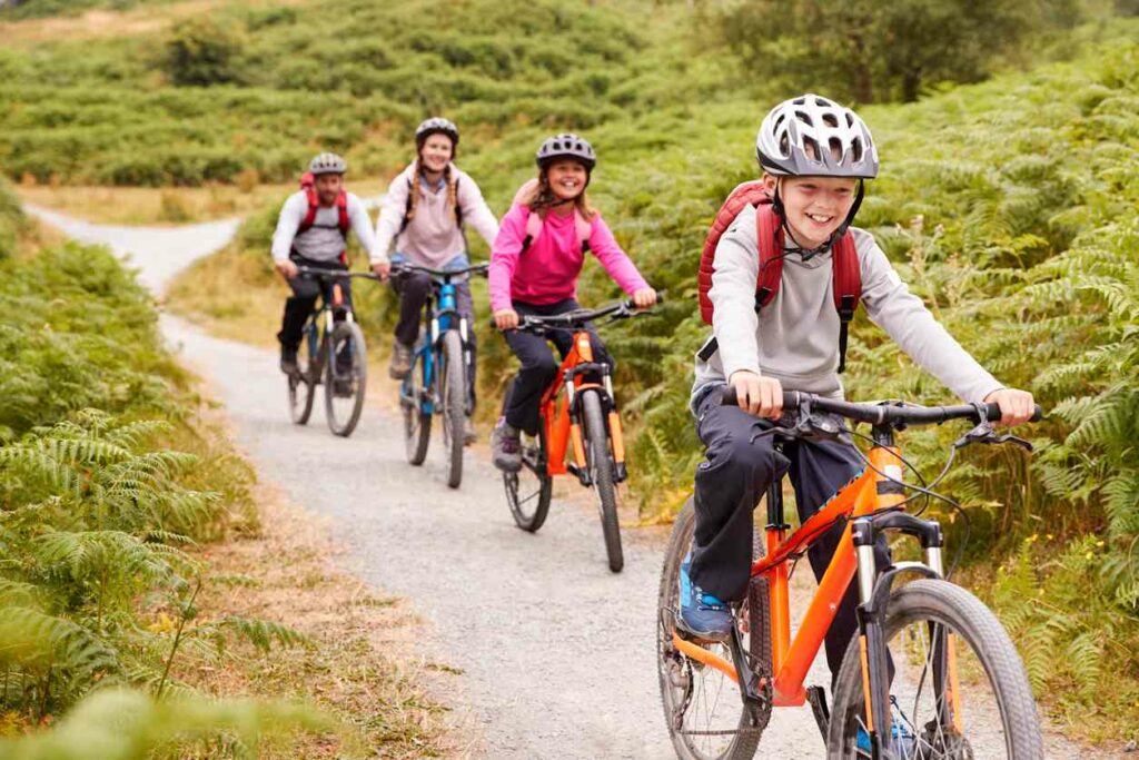 A family of four rides their bikes on a gravel road surrounded by green brush and grass