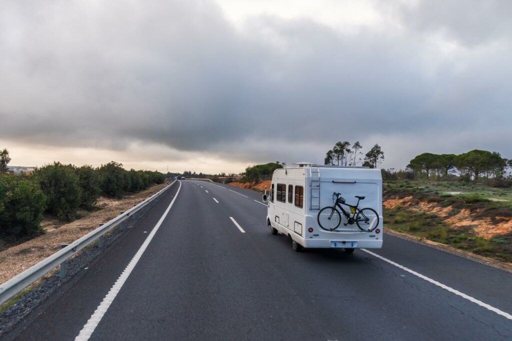 A white RV with a bicycle attached to the back is driving away down a two lane empty road