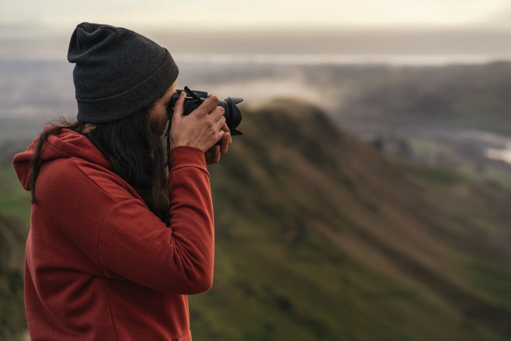 A woman in a red sweatshirt and black beanie uses a camera to take a photo. A mountain scene can be seen behind her