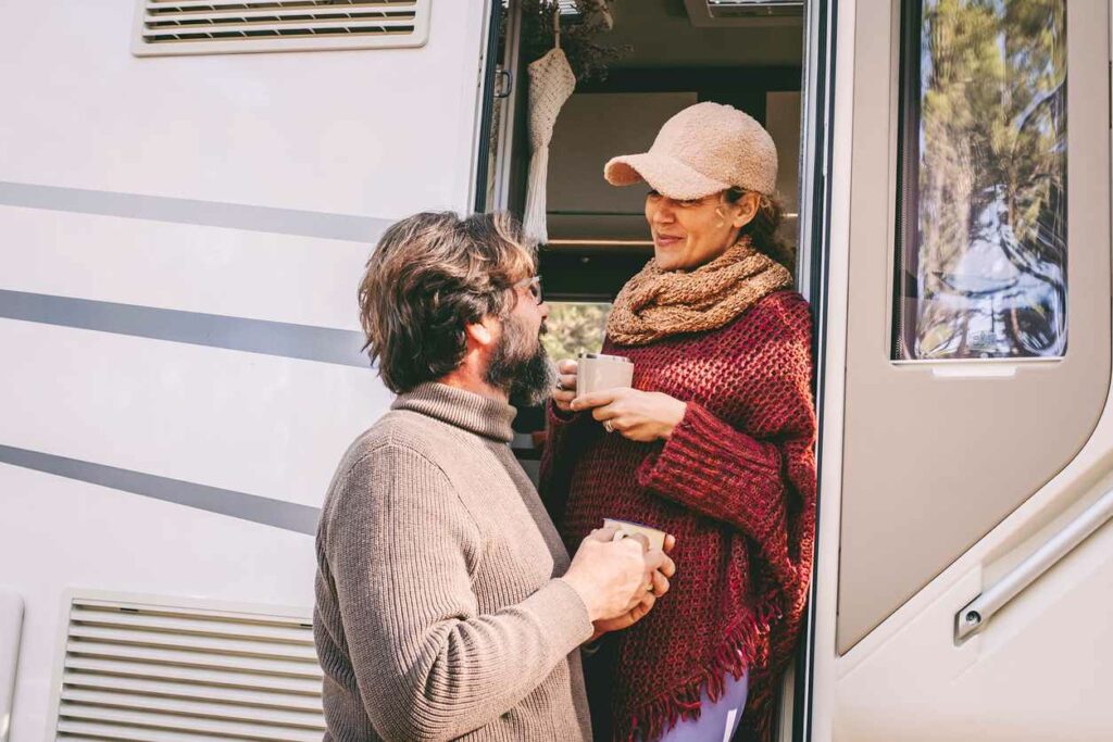 A man and woman stand in the doorway of an RV wearing fall clothing and drinking coffee