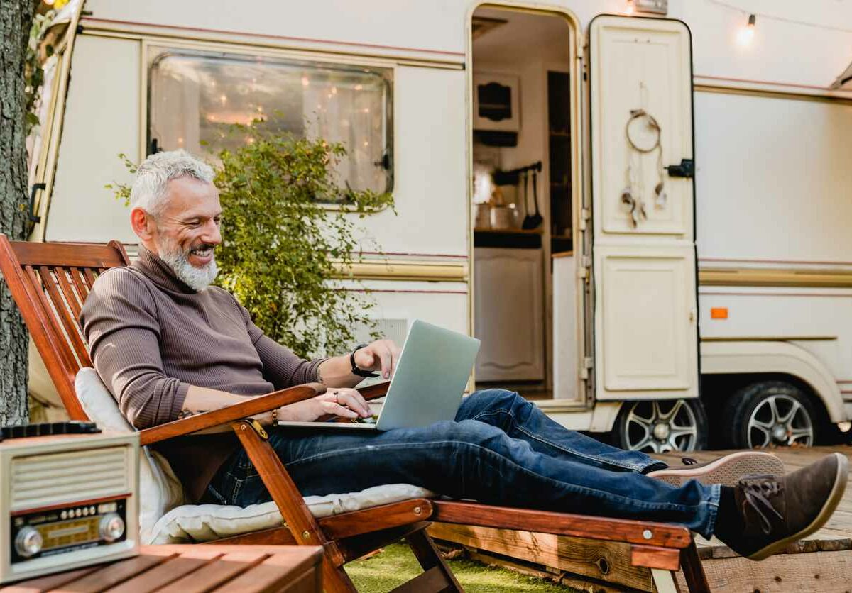 A man with white hair and a white beard relaxes on a wooden chair outside of a white RV with the door open