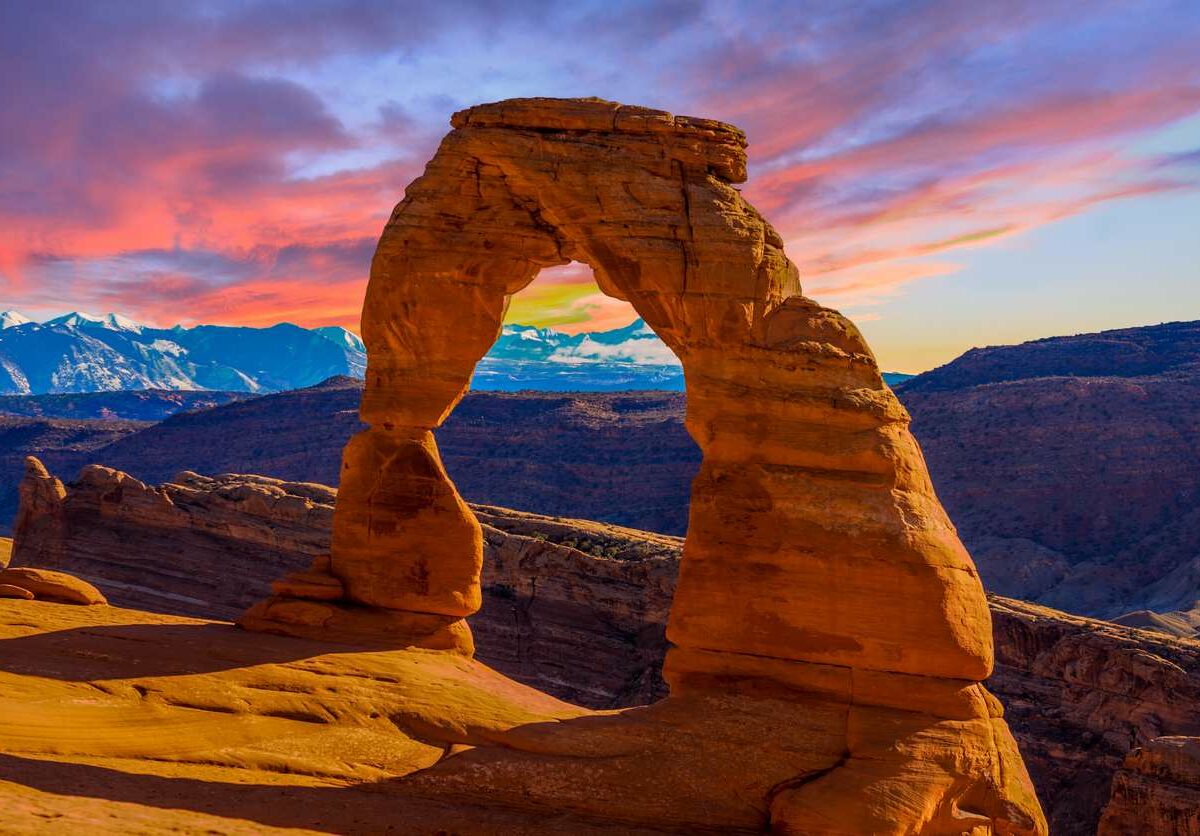 The sun sets over an orange stone arch at Arches National Park, with a pink, orange, and blue sky in the background