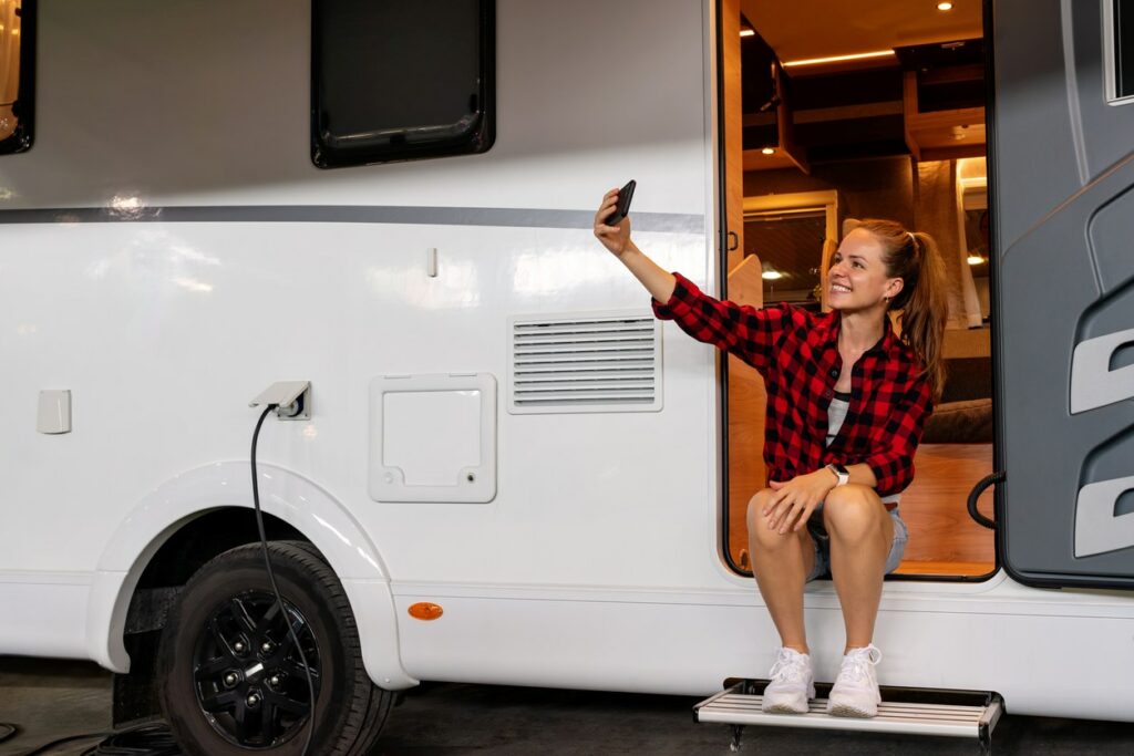 A young woman taking a photo on her phone while sitting at the door of her RV.