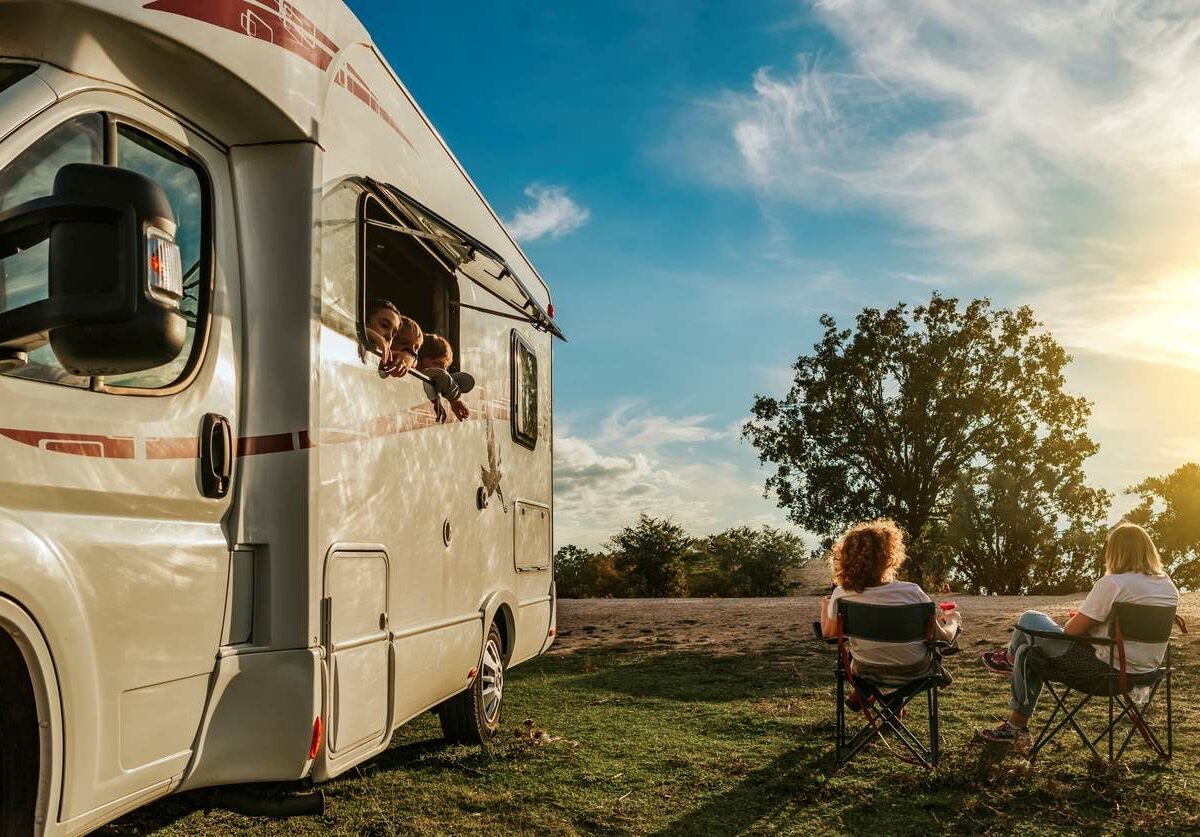 Children leaning out of an RV window watching the sunset while their mothers drink coffee in fold-up chairs.