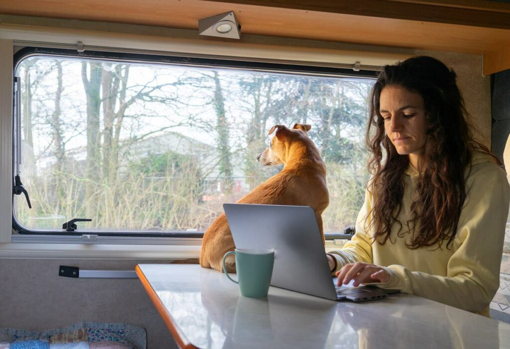 A woman with a dog working on a laptop inside an RV.