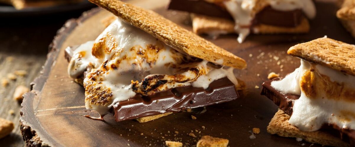 Campfire s’mores are sitting and melting on a wooden platter and table.