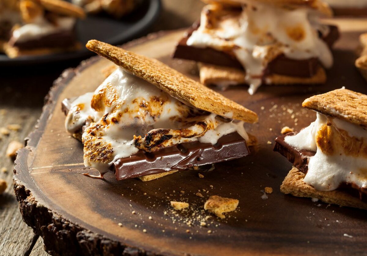 Campfire s’mores are sitting and melting on a wooden platter and table.