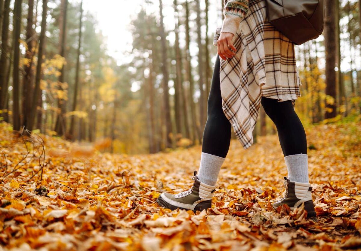 Woman’s legs in hiking boots traveling a forested path covered with fallen leaves.