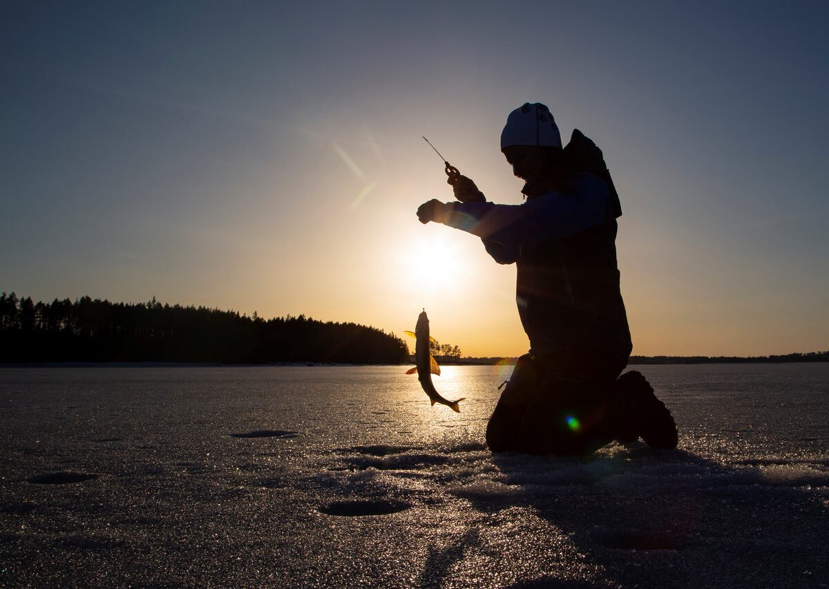 A silhouette of a woman ice fishing on a frozen lake during sunset.