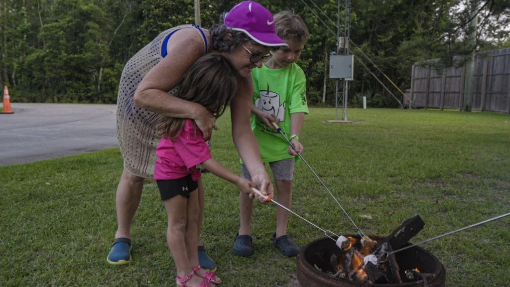 A grandmother helps her grand daughter roast a marshmallow along side another little boy over a firepit at Lakeside RV Resort by RJourney.