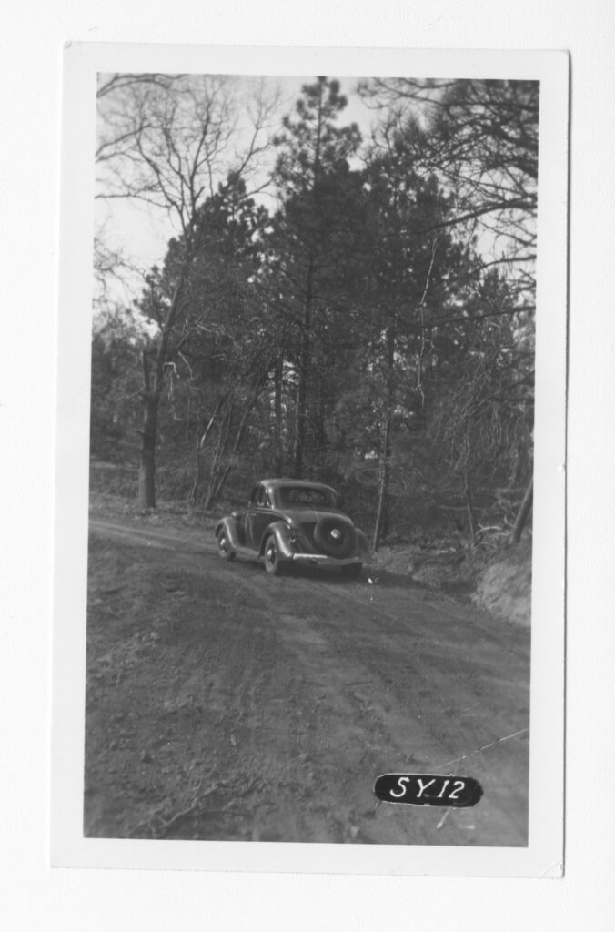 An old black car turns left on an unpaved road.