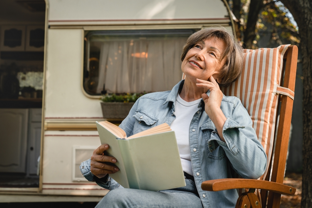 An older woman contemplates the words in a book in front of her RV.