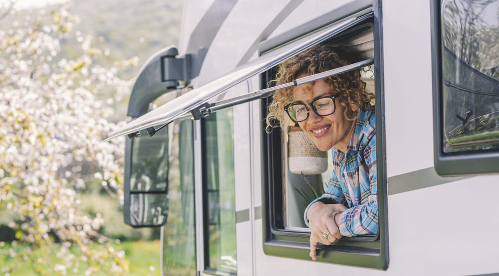 A smiling young woman in glasses looks out the open window of an RV out into nature.